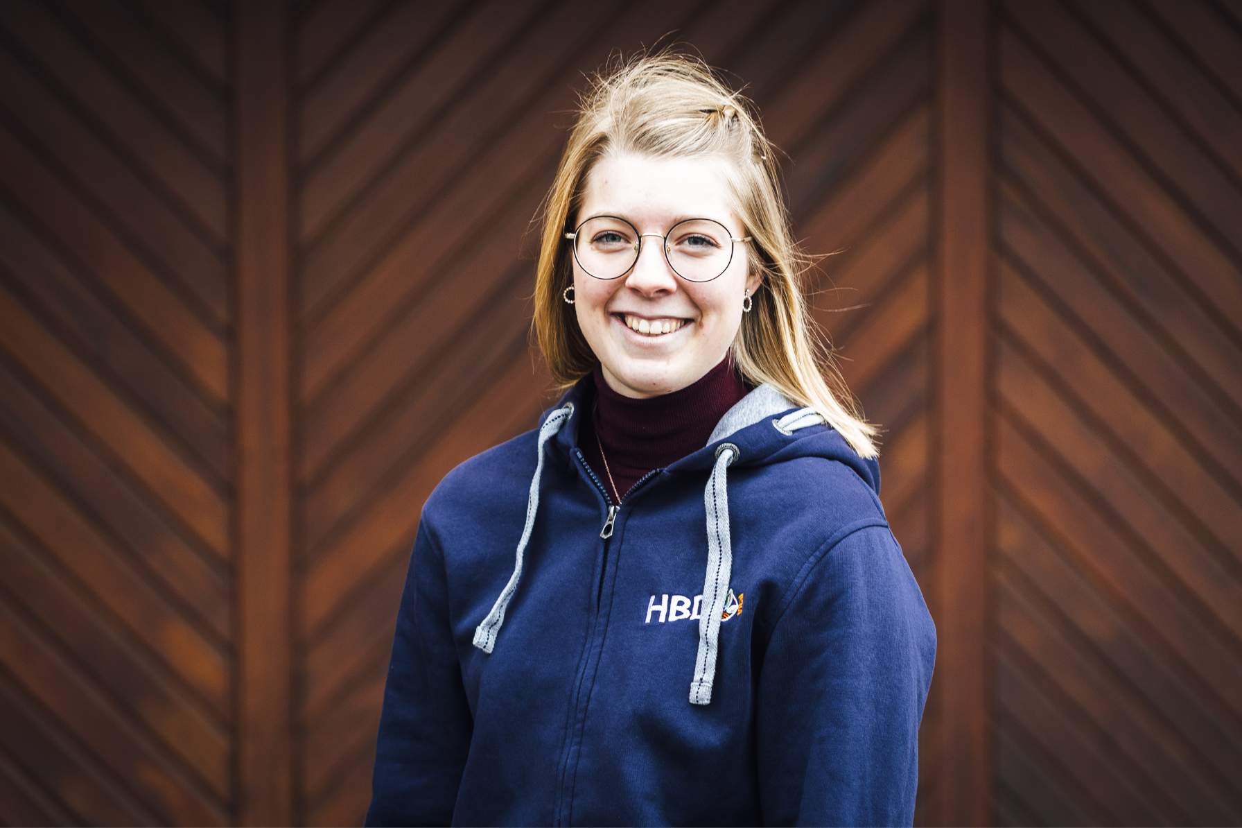 Emily Beifuss Pensionsmanagement Managerin der Pension am Hohberghof, Emily Beifuss ist auf dem Bild zu sehen. Sie steht vor einem dunkelbraunen Tor und trägt einen blauen HBD-Hoodie