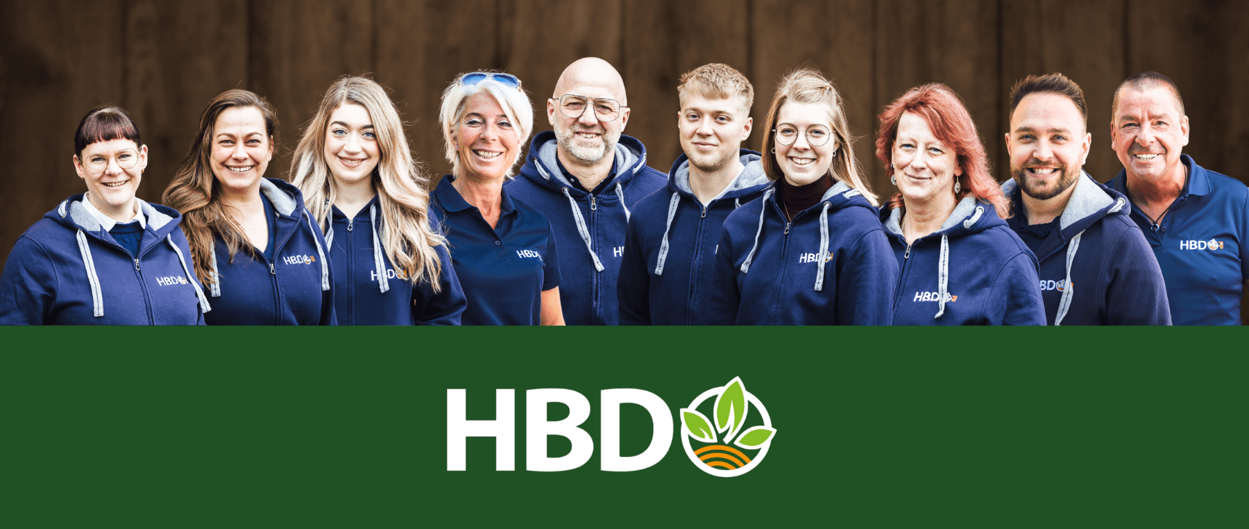 Das Team von HBD Es ist das komplette HBD Team mit all seinen Mitarbeitern vor einem braunen Holztor zu sehen.