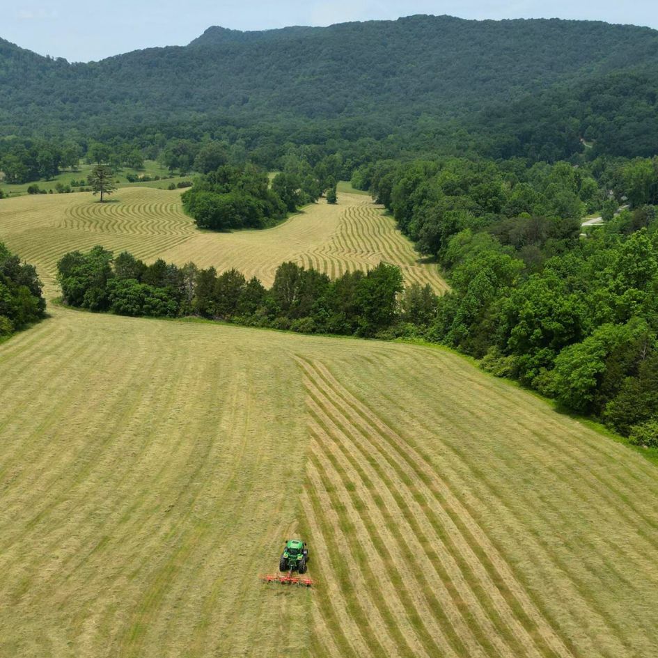 Bild zu Blogbeitrag: Grünlandpflege für Pferdeweiden Foto von Feldern auf denen ein Schlepper fährt und die Weide bearbeitet. Im Hintergund sind Berge mit vielen grünen Bäumen.