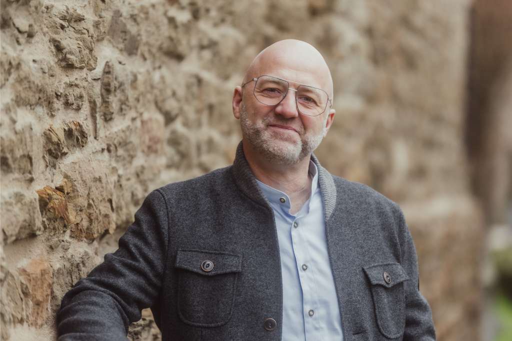 Coaching Heiner Beifuss Consulting Agrarsektor Porträt von Heiner Beifuss mit Brille und grauem Jacket, der an einer Steinmauer lehnt