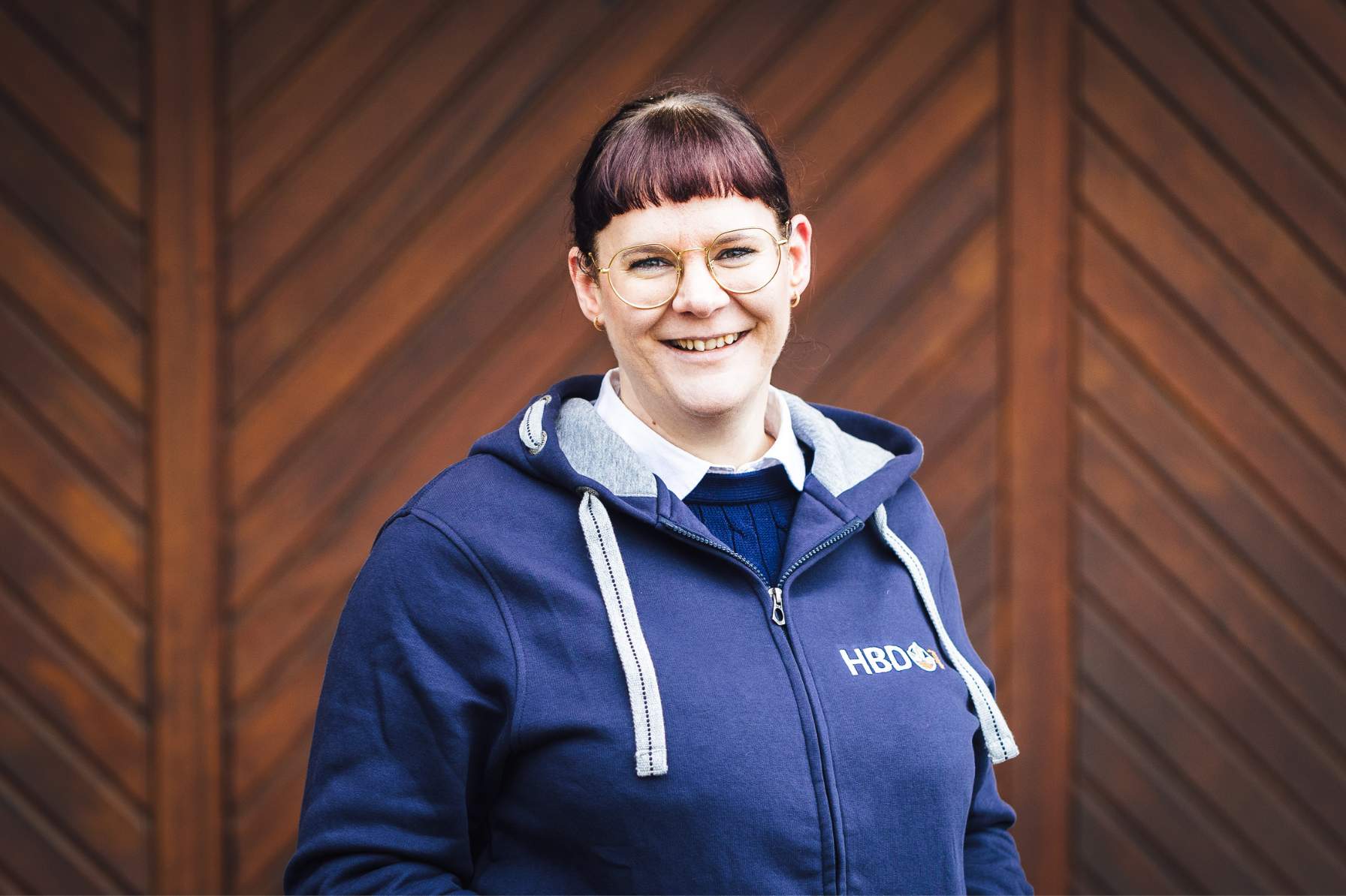 Marketing Frau Schoenemann Frau Schoenemann aus der Marketingabteilung steht vor einem braunen Tor und trägt einen blauen HBD-Hoodie