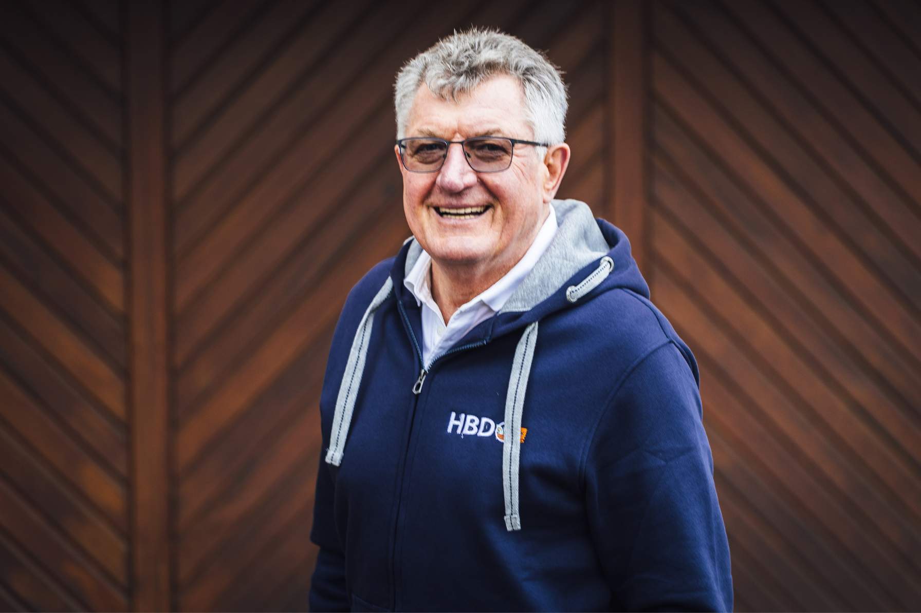 Facility Manager Herr Wickenheißer Herr Wickenheißer der Facility-Manager von HBD steht vor einem dunkelbraunen Tor und trägt einen blauen HBd-Hoodie