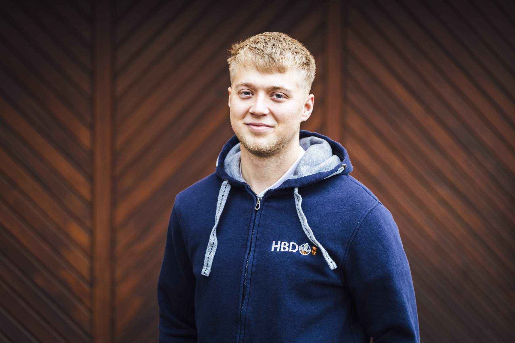 Social-Media Assitent Hendrik Beifuss Hendrik Beifuss der bei HBD als Social-Media-Assistent arbeitet, steht vor einem dunkelbraunen Tor und trägt einen blauen HBD-Hoodie