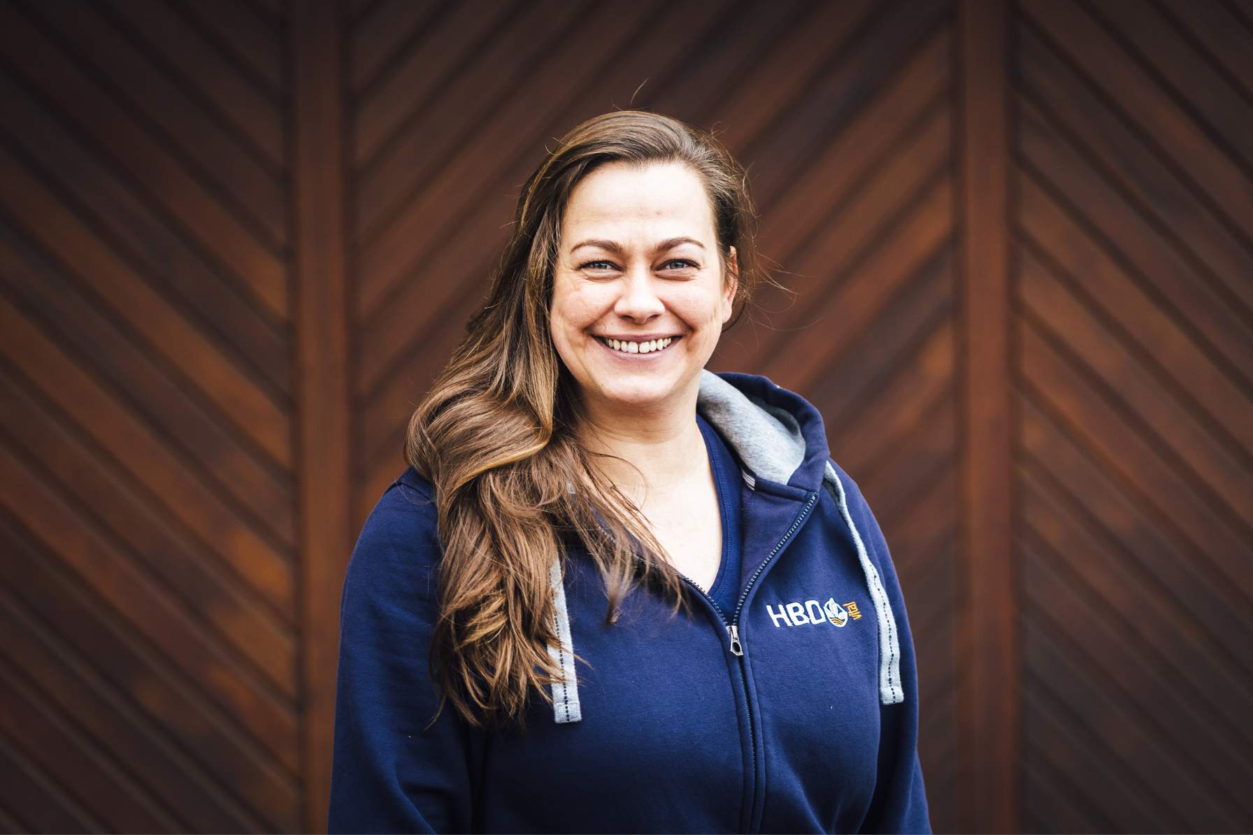 Marketing Frau Lisken Frau Lisken aus der Marketingabteilung steht vor einem braunen Tor und trägt einen blauen HBD-Hoodie