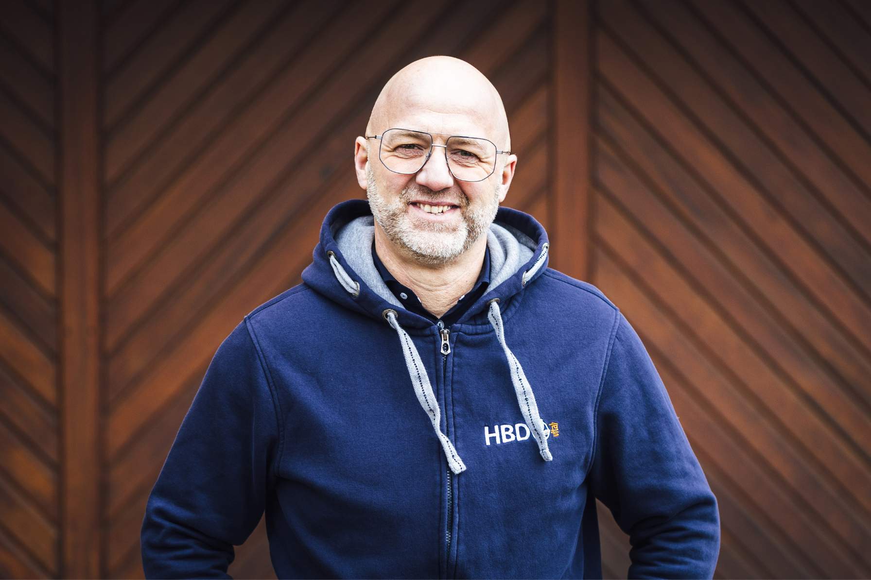 Geschäftsführer Heiner Beifuss Es ist der Geschäftsinhaber Heiner Beifuss zu sehen. Dieser trägt eine Brille und ein einen blauen HBD Hoodie und steht vor einem dunkelbraunen Tor