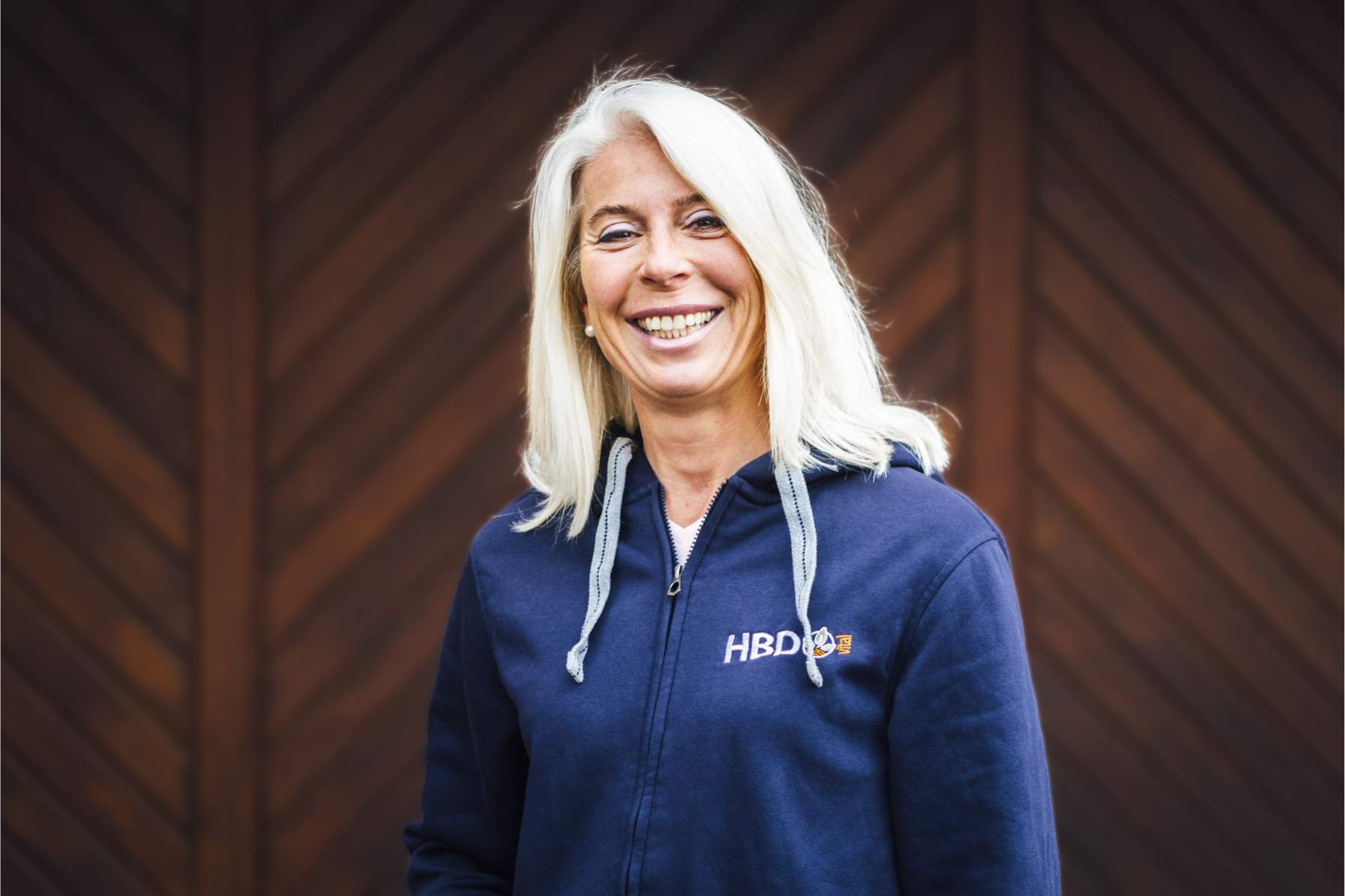 Anja Beifuss Geschäftsführerin HBD Vital Anja Beifuss steht vor einem dunkelbraunen Tor und trägt einen blauen HBD-Hoodie