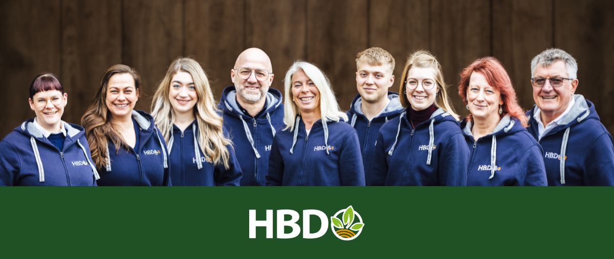 Das Team von HBD Es ist das komplette HBD Team mit all seinen Mitarbeitern vor einem braunen Holztor zu sehen.